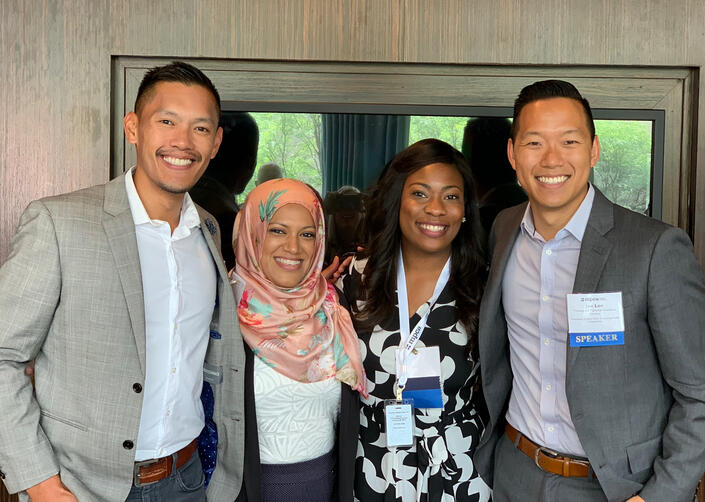Speaking Engagement Four professionally dressed people stand close together and smile for a photo at a conference, with one person wearing a floral headscarf and another wearing a name badge labeled “Speaker.”