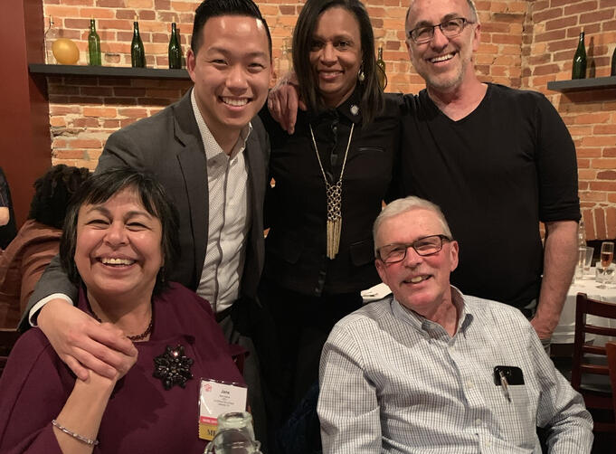 Mentors A group of five adults pose together at a restaurant with exposed brick walls and bottles on shelves in the background, smiling and leaning in close around a table during a social gathering.
