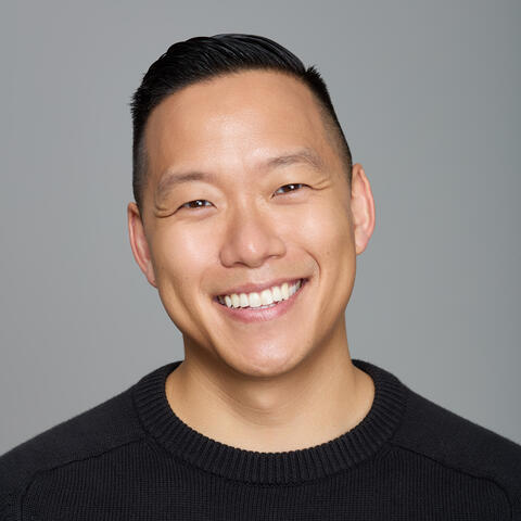Professional headshot of a person in a dark sweater, shown from the shoulders up against a gray background within a teal circular frame.